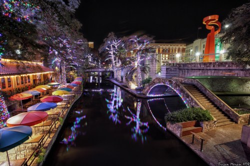 A vibrant night scene of a canal with colorful lights, umbrellas, and a modern sculpture in the background.