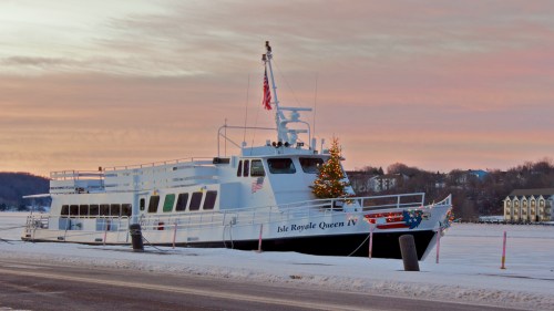 A festive boat named Isle Royale Queen IV docked in snow, decorated with a Christmas tree against a sunset sky.