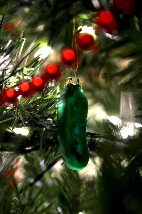 A green pickle ornament hanging on a Christmas tree, surrounded by red garland and soft white lights.