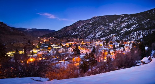 A scenic view of a snowy mountain town at dusk, with lights twinkling in the valley and mountains in the background.