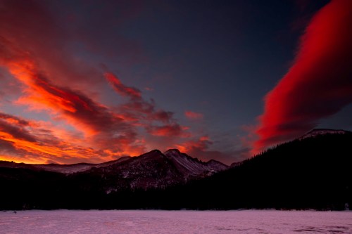 A dramatic sunset paints the sky in red and orange hues over snow-covered mountains and a dark forest silhouette.