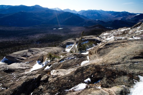 A panoramic view of rugged mountains and valleys, with rocky terrain and patches of snow under a clear blue sky.