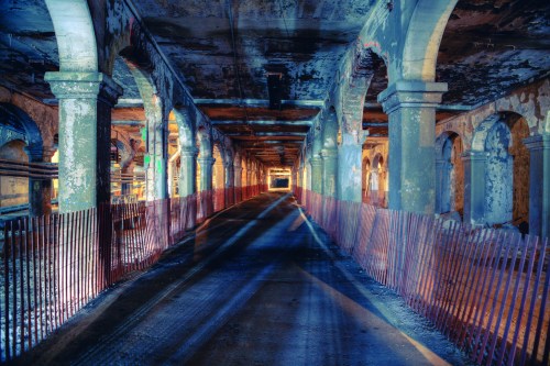 An abandoned underground tunnel with arched columns and a dimly lit pathway, featuring a fence along the sides.