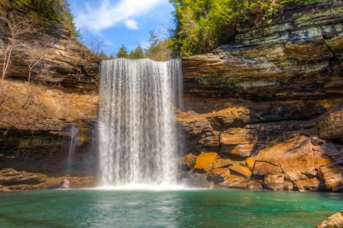 A stunning waterfall cascades over rocky cliffs into a clear pool, surrounded by lush greenery and a blue sky.