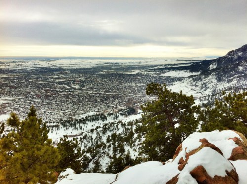 A snowy landscape view from a mountain, overlooking a valley and distant hills under a cloudy sky.