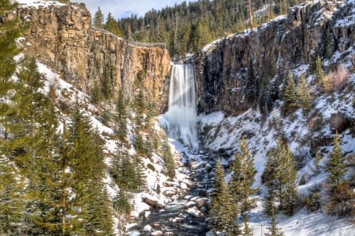 A stunning winter scene featuring a waterfall cascading down rocky cliffs, surrounded by snow-covered trees and a river.