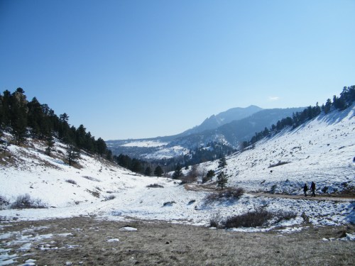 A snowy landscape with mountains in the background and a clear blue sky, featuring a winding path and two hikers.