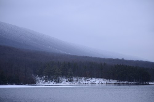 A serene winter landscape featuring a snowy mountain, a calm lake, and a forested shoreline under a gray sky.