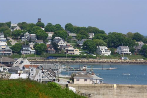 A coastal view of a small town with houses on a hillside and a marina in the foreground under a clear blue sky.
