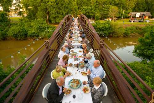 A long table set for a dinner party on a bridge, surrounded by greenery and a river, with guests enjoying their meal.