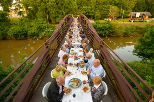A long table set for a dinner party on a bridge, surrounded by greenery and a river, with guests enjoying their meal.
