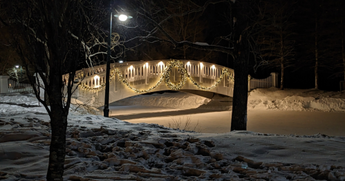 A snow-covered bridge at night, adorned with festive lights and surrounded by trees in a winter landscape.