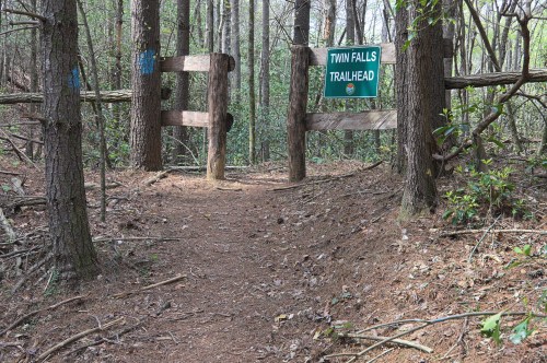 A wooden gate marks the entrance to the Twin Falls Trailhead, surrounded by trees and a dirt path.