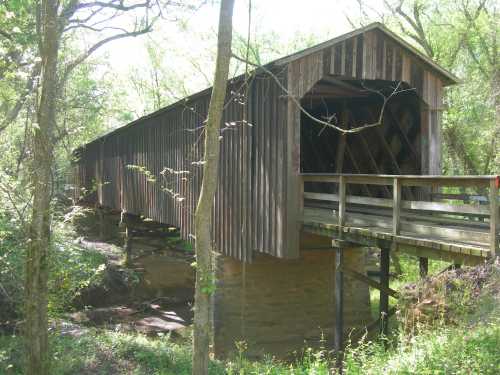 A wooden covered bridge stands over a stream, surrounded by lush greenery and trees. Sunlight filters through the leaves.