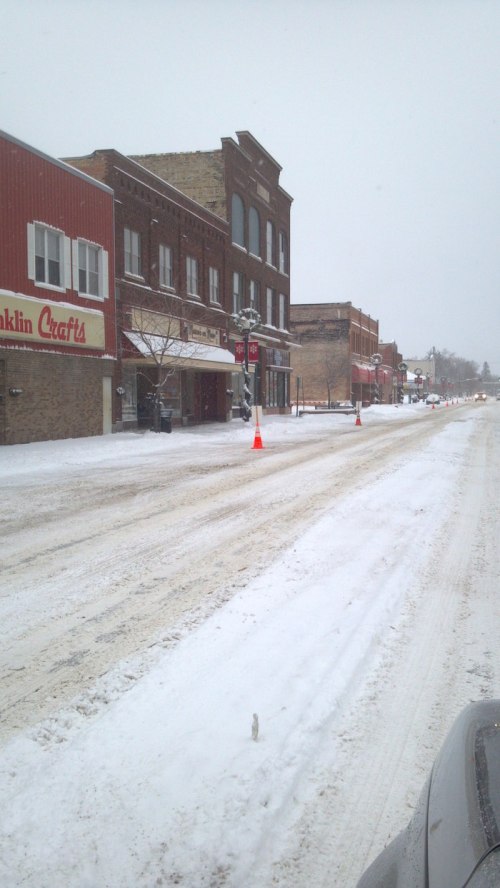 Snow-covered street in a small town, with shops lining the road and traffic cones marking the way.