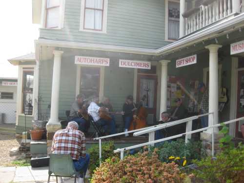 A group of musicians playing instruments on a porch, with a gift shop and music signs in the background.