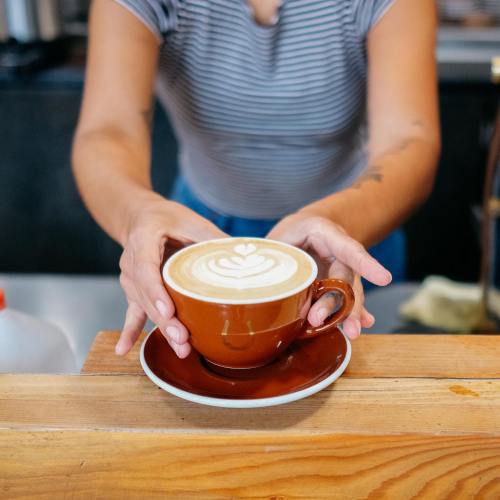 A person holds a brown cup of latte art over a wooden counter, with a small plate underneath.