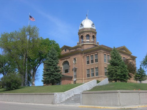 Historic brick building with a dome, surrounded by trees and a clear blue sky, featuring an American flag.
