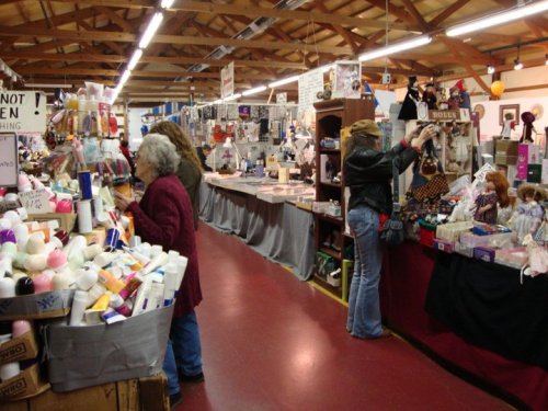 A bustling indoor market with vendors selling various items, including dolls and beauty products, and shoppers browsing.