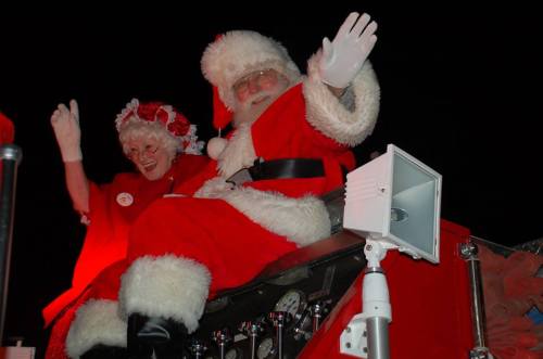 Santa Claus and a woman in festive attire wave from a decorated sleigh, surrounded by festive lights.
