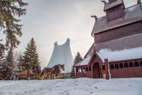 A snowy landscape featuring a wooden building and a horse-drawn carriage, with trees and a large white structure in the background.
