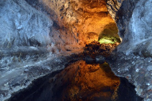 A dimly lit cave with colorful rock formations and a reflective pool of water on the ground.