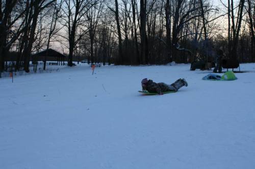 A child sleds down a snowy hill, surrounded by trees and a few tents in the background during dusk.