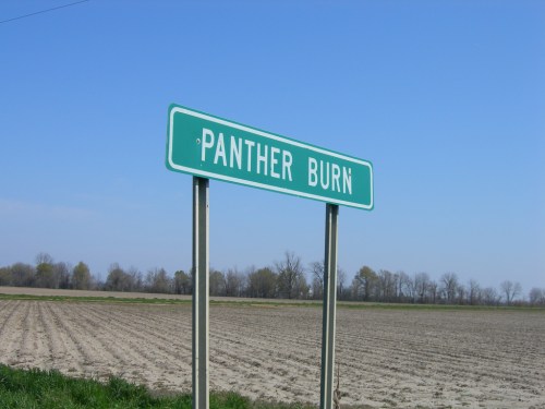 A green road sign reading "Panther Burn" stands beside a plowed field under a clear blue sky.