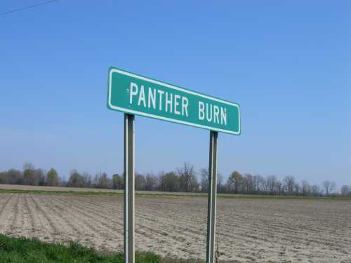 A green road sign reading "Panther Burn" stands beside a plowed field under a clear blue sky.