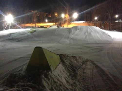 Snow-covered terrain park at night, featuring ramps and obstacles illuminated by bright lights.
