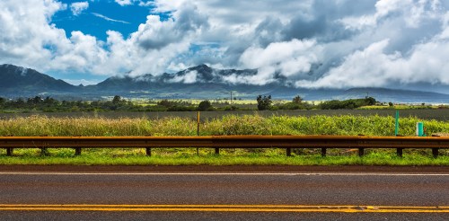 A scenic view of a road with lush green fields and mountains under a cloudy sky in the background.