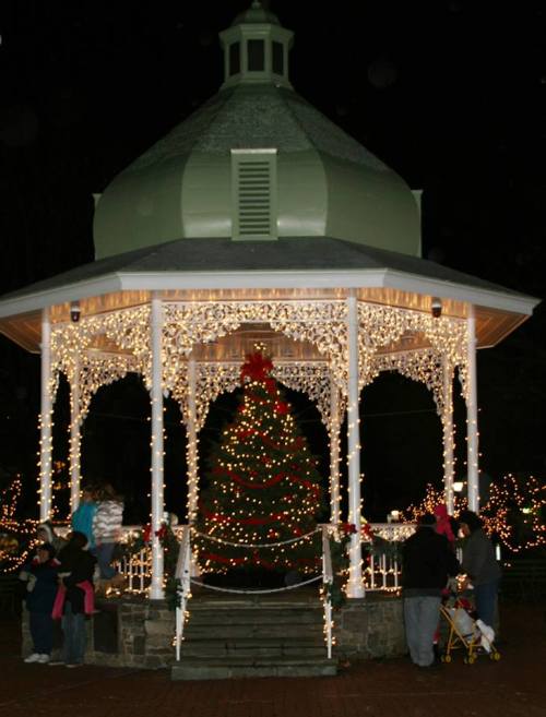 A festive gazebo adorned with lights and a Christmas tree, surrounded by people enjoying the holiday atmosphere at night.