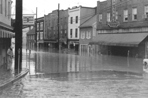 A flooded street with submerged buildings and parked cars, reflecting the water's surface in a black-and-white photo.