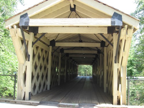 A wooden covered bridge with a lattice design, leading into a green, tree-lined area.