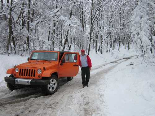 A man stands beside an orange Jeep in a snowy forest, with trees covered in white snow lining the road.