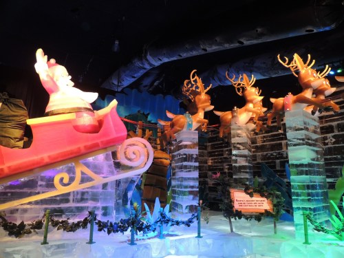 A colorful ice sculpture of Santa in a sleigh with reindeer, surrounded by festive decorations and icy structures.