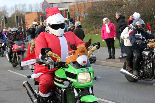 A motorcyclist in a Santa suit rides a green bike, with a reindeer decoration on the front, during a festive parade.
