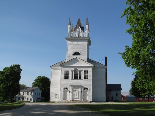 A white church with a tall steeple, surrounded by green trees and a clear blue sky.