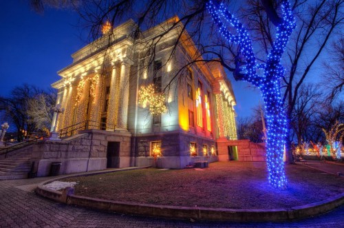 A beautifully lit building adorned with holiday lights, surrounded by trees glowing with blue and warm lights at dusk.