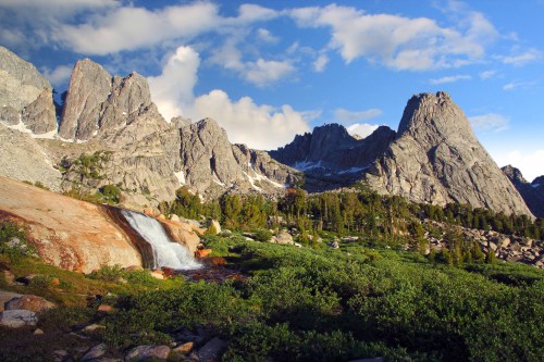 A scenic view of rugged mountains with a waterfall and lush greenery under a partly cloudy sky.