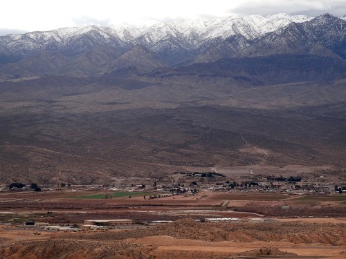 A panoramic view of a valley with a small town, surrounded by mountains capped with snow under a cloudy sky.