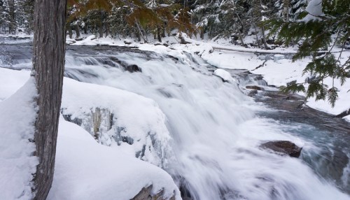 A snowy landscape featuring a flowing river with cascading water and trees in a winter setting.