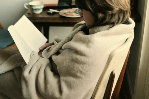 A person wrapped in a cozy blanket sits in a chair, reading a book with a plate and cup nearby.