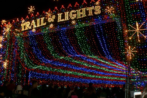 Colorful holiday lights create a vibrant display at the "Trail of Lights" entrance, with a crowd gathered below.