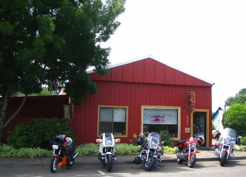 A red building with a sign, surrounded by greenery, featuring several parked motorcycles in front.