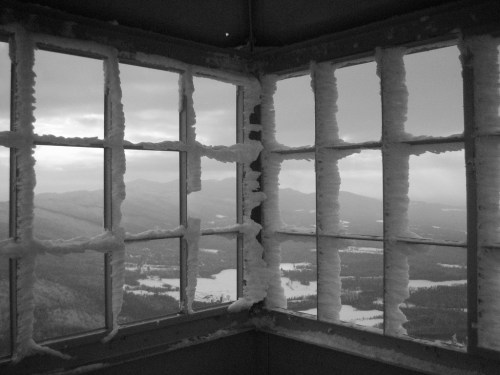 Black and white image of a window with frost, overlooking a snowy mountain landscape.