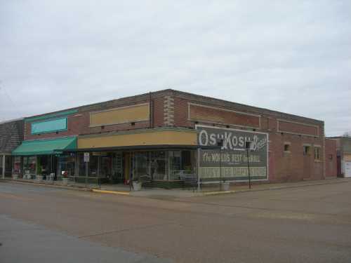 A brick storefront with a yellow awning and vintage signage, located on a quiet street under a cloudy sky.