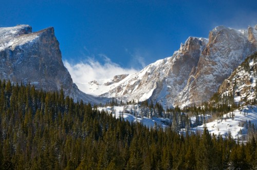 Snow-capped mountains rise above a dense forest under a clear blue sky, with wispy clouds drifting by.