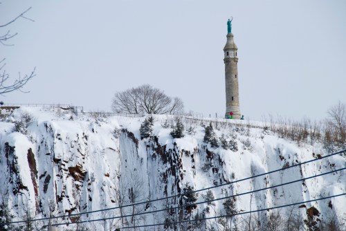 A snowy cliff with a tall monument and statue on top, surrounded by trees and a clear winter sky.
