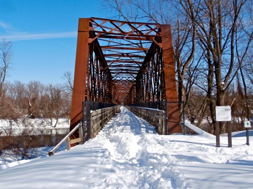 A snow-covered bridge with a metal frame spans a river, surrounded by bare trees under a clear blue sky.